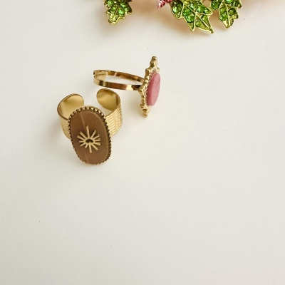 Dois anéis dourados com detalhes em pedra castanha e rosa, folhas verdes ao fundo.