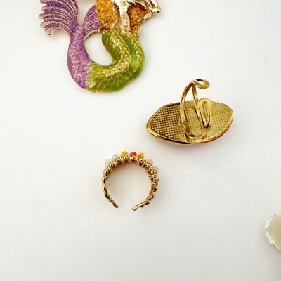 Anel dourado com pedras, broche dourado em forma de chinelo e broche sereia colorida em fundo branco.