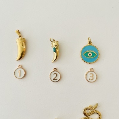 Seis pendentes dourados e numerados sobre fundo branco, incluindo chifres, um olho azul, cobra, flor e sol.