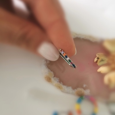 Anel fino prateado com pedras coloridas segurado por dedo com unha branca sobre fundo de pedra rosa