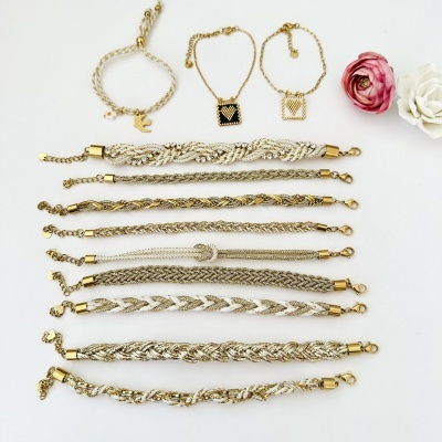 Pulseiras douradas e brancas com pendentes e malhas entrelaçadas sobre fundo branco e flores artificiais.