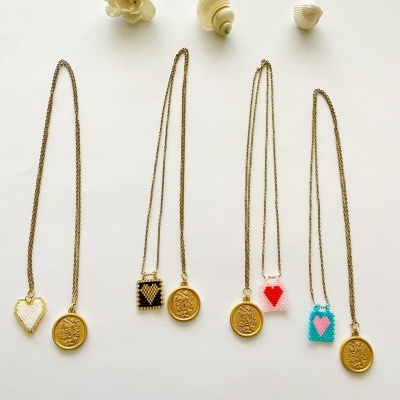Quatro colares dourados com pendentes redondos e pequenos quadrados com corações coloridos numa superfície branca com conchas e flor decorativa