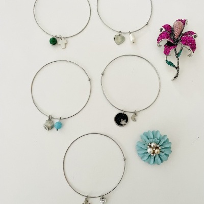 Coleção de pulseiras com pendentes variados e dois broches florais coloridos sobre fundo branco.