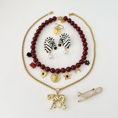 Bijuterias diversas com temas de animais e pedras falsas, na cor dourada, vermelha e detalhes em preto e branco, sobre fundo branco.