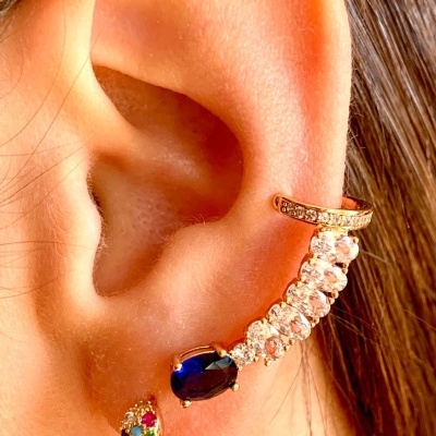 Orelha com três brincos dourados com pedras brilhantes transparentes, azul e coloridas.