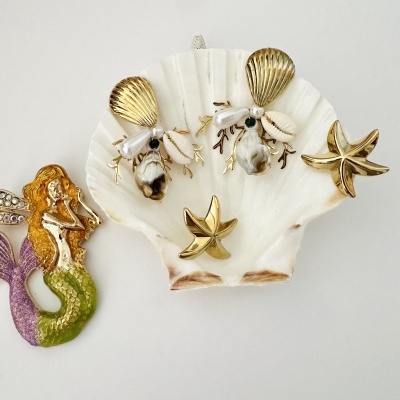 Brincos decorados com conchas e pérolas sobre concha branca com figura de sereia e estrela-do-mar dourada