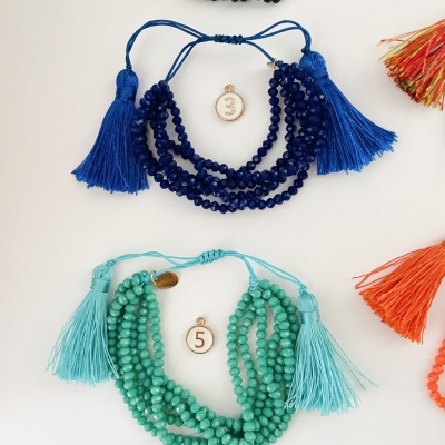 Pulseiras com contas de pedra azul escuro e verde água com tassels e etiquetas numéricas, em fundo branco com franjas coloridas.