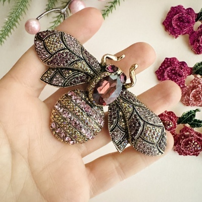 Broche em forma de abelha com pedras rosa e douradas numa mão com flores rosas ao fundo