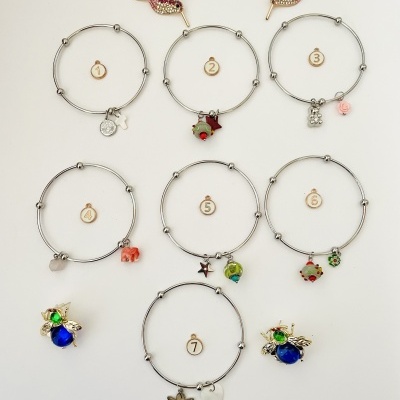 Oito pulseiras prateadas com pendentes coloridos dispostas sobre fundo branco, com dois pássaros rosa no topo.