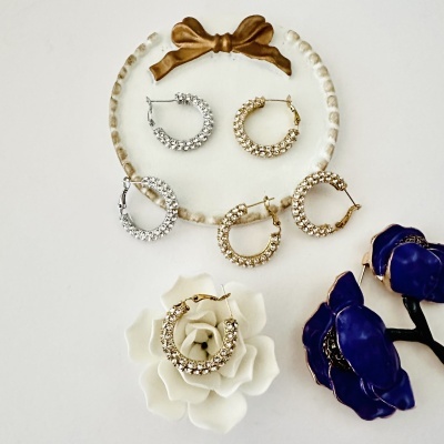 Brincos de argola dourados e prateados com pedras brilhantes sobre superfície branca com laço dourado e flores decorativas