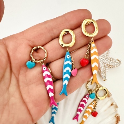 Brincos coloridos em forma de peixe com padrões brancos, feitos em metal dourado com corações pendurados numa mão