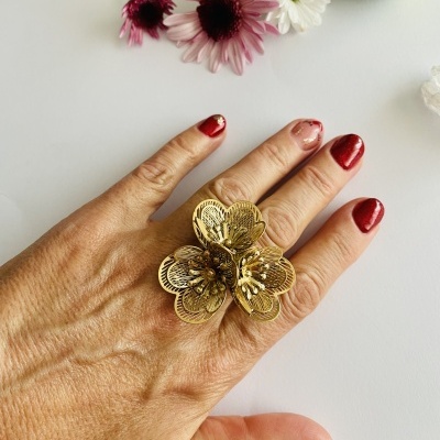 Anel dourado em forma de flor numa mão com unhas vinho, flores naturais ao fundo