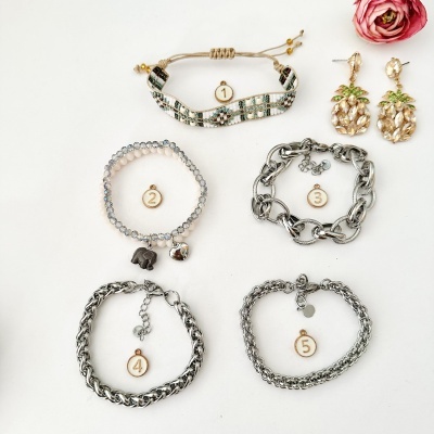 Cinco pulseiras numeradas e brincos de abacaxi dourados sobre fundo branco com flor rosa no canto