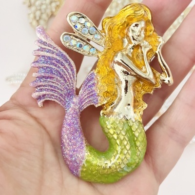 Broche de sereia colorido com cauda brilhante, cabelo amarelo e asas com pedrinhas azuis na mão