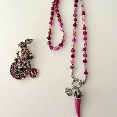 Colar com contas rosa/vermelho e pendente de chifre rosa com detalhe prateado, mais broche de coelho em bicicleta com pedraria rosa, tudo sobre fundo branco