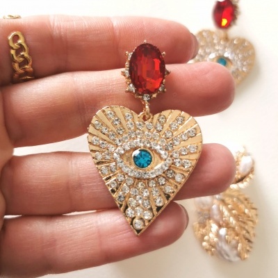 Brincos dourados em forma de coração com pedras brilhantes e pedra azul no centro em forma de olho, pendurados numa pedra vermelha oval