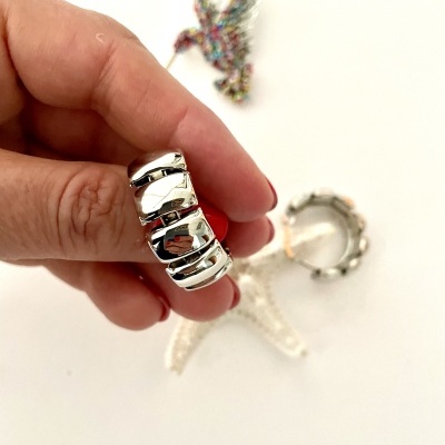 Brinco de argola prateada segurado por mão com unhas vermelhas, fundo branco com broche de pássaro colorido e outro brinco