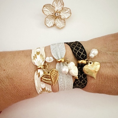 Pulso com três pulseiras elásticas douradas, prata e pretas com pingentes de coração e pérola