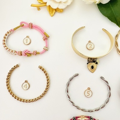 Cinco pulseiras diversas numeradas com pingentes, sobre superfície branca com flores e folhas ao fundo.
