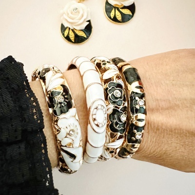 Braceletes dourados com detalhes em preto e branco num pulso e flores brancas ao fundo