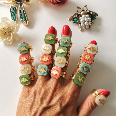 Mão com anéis coloridos com símbolos de abelha, unhas vermelhas e brancas, flores artificiais e broches no fundo.