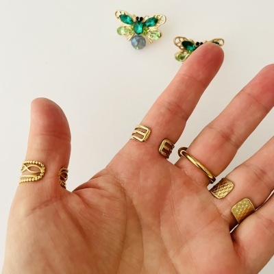 Mão com cinco anéis dourados e dois brincos de borboleta com pedras verdes e azuladas sobre fundo branco.