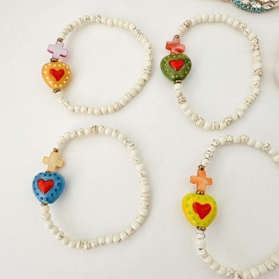 Pulseiras elásticas com contas brancas e peças centrais de coração e cruz em cores diversas.
