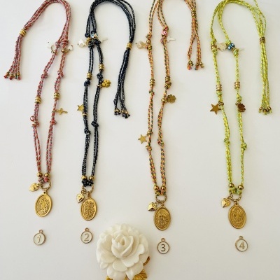 Quatro colares coloridos com contas, medalhas douradas e pendentes, numerados de 1 a 4, sobre fundo branco, com flor branca decorativa