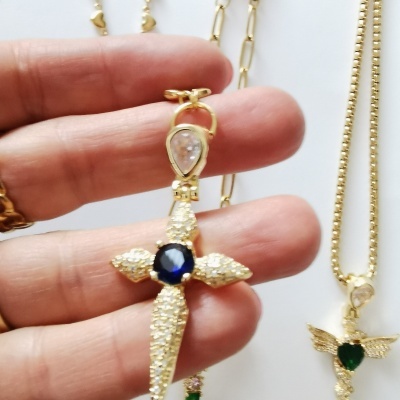 Colares dourados com pendentes em cruz e anjo, com pedras brilhantes e azuis, segurados por mão.