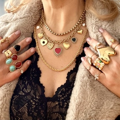 Conjunto de colares e anéis dourados com pedras e pendentes de coração sobre blusa preta de renda e casaco bege de pelo.