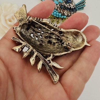 Broche em forma de pássaro com pedras azuis e douradas, pino de segurança na parte de trás, segurado numa mão.