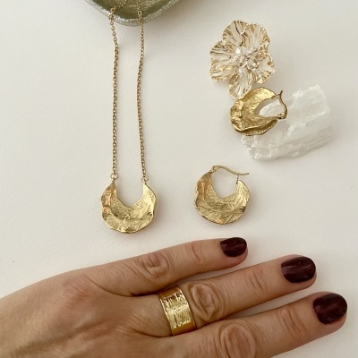 Conjunto de joias douradas texturizadas com colar, brincos e anel numa mão com unhas pintadas de escuro.