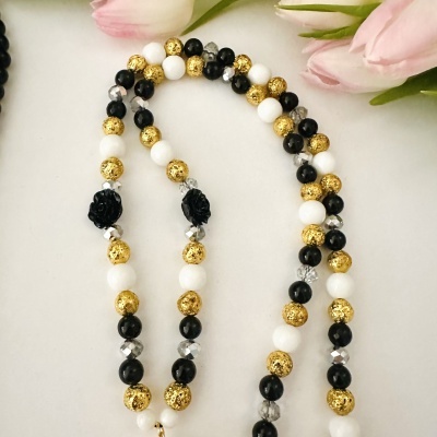 Colar com contas pretas, brancas, douradas, transparentes e duas contas pretas em formato de rosa sobre fundo branco e flores cor-de-rosa