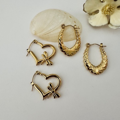 Quatro brincos dourados em aro e coração com decoração de asas e libélula, sobre superfície branca com concha e flor decorativa.