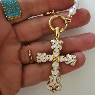 Colar com cruz branca e dourada segurado por mão com anel azul e dourado