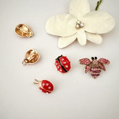 Brincos joaninha vermelha e abelha rosa com pedras brilhantes, em superfície branca com flor branca