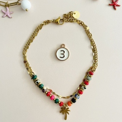 Pulseira dourada com contas coloridas e pendente de palmeira sobre fundo bege