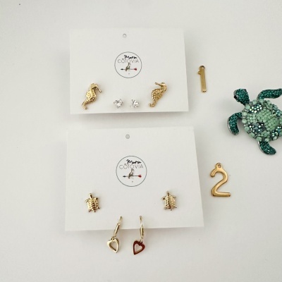Dois conjuntos de brincos dourados em cartões com logótipo Maria COTOVIA e acessórios decorativos ao lado.