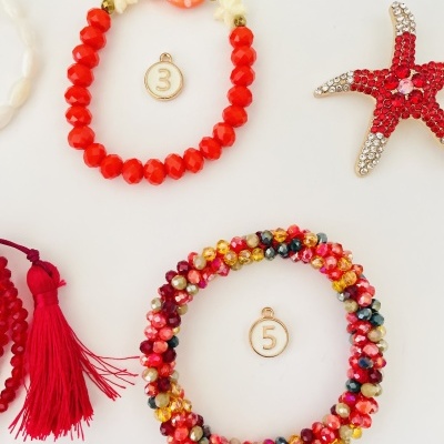 Dois braceletes de contas vermelhas e multicoloridas, estrela do mar vermelha com cristais e fio com borla vermelha sobre fundo branco.