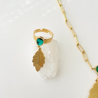 Anel dourado com pedra verde e pendente de folha, sobre cristal branco, com corrente dourada visível