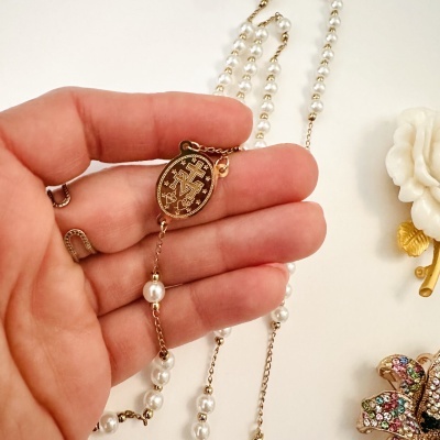 Colar com contas brancas e pendente oval dourado com símbolo gravado segurado por mão com anéis dourados