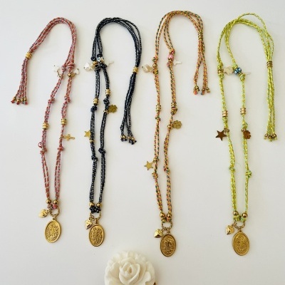 Quatro colares trançados coloridos com medalhões dourados e pendentes, sobre fundo branco e rosa decorativa