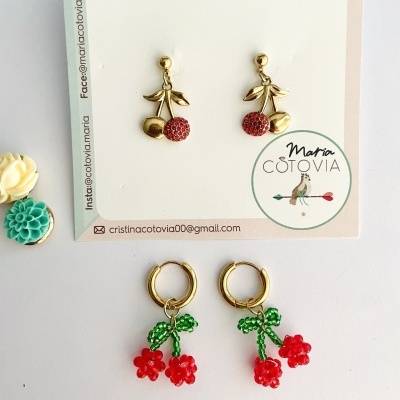 Dois pares de brincos com design de cerejas, um dourado e outro com contas vermelhas e verdes, sobre fundo branco com etiquetas e flores