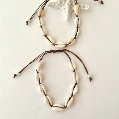 Duas pulseiras com conchas brancas e cordão castanho sobre fundo branco com estrela-do-mar decorativa