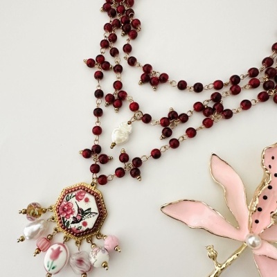 Colar com contas vermelhas e pendente decorativo com desenho floral e pássaro, e broche flor rosa com detalhes dourados.