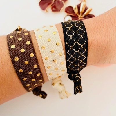 Pulseiras de tecido castanho, creme e preto com padrões dourados e brincos ao fundo