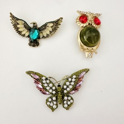 Três broches decorativos em forma de águia, coruja e borboleta com pedras coloridas sobre fundo branco.