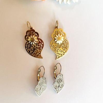 Quatro pares de brincos de metal com design floral em dourado, castanho e prateado, dispostos em superfície branca com flores artificiais ao fundo