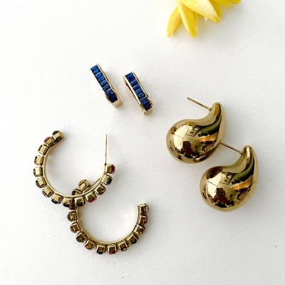 Três pares de brincos dourados com diferentes estilos e pedras coloridas, sobre fundo branco com flor amarela