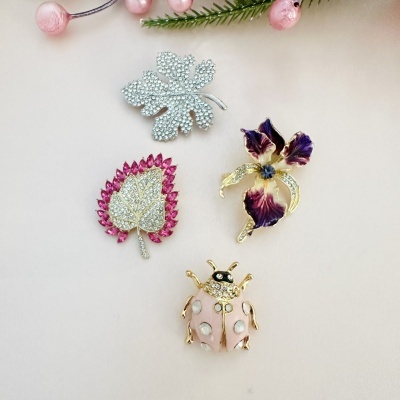 Quatro broches decorativos em formas naturais com pedras e esmalte, em fundo bege com ramos e bagas cor-de-rosa.